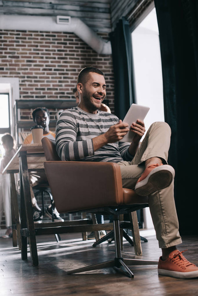 alegre hombre de negocios usando tableta digital mientras está sentado en sillón cerca de un colega afroamericano
 - Foto, Imagen