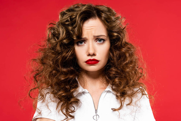 attractive upset curly girl looking at camera, isolated on red - Фото, изображение