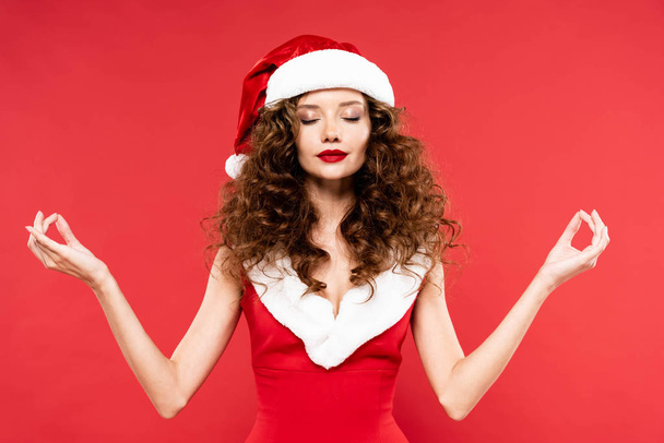 calm curly girl posing in santa costume with gyan mudra signs, isolated on red - 写真・画像