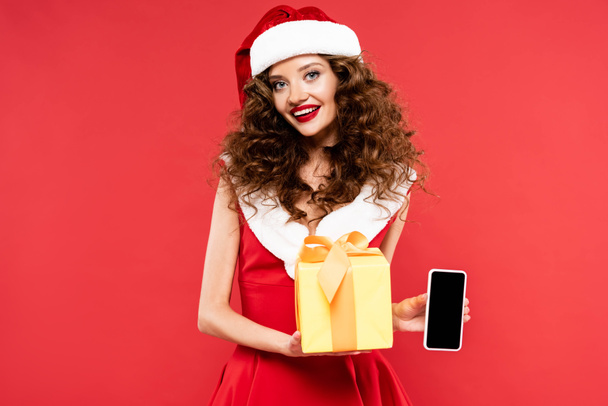 attractive smiling girl in santa costume holding christmas gift and smartphone with blank screen, isolated on red - Fotografie, Obrázek