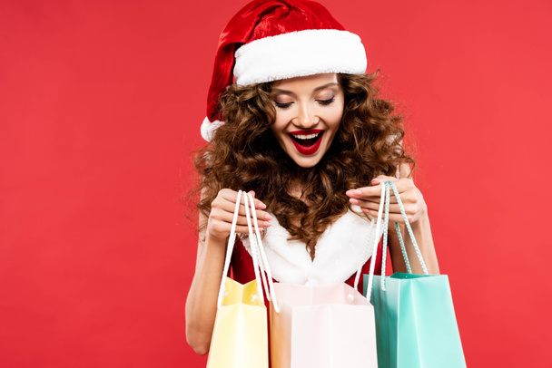 excited curly girl in santa costume posing with shopping bags, isolated on red - Foto, immagini