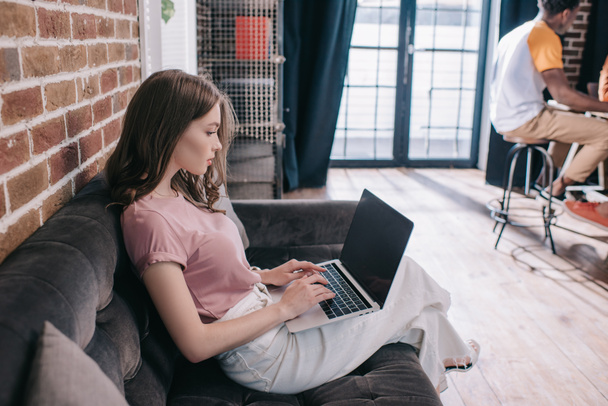 young, thoughtful businesswoman using laptop while sitting on sofa in office - Foto, Bild