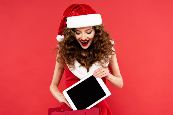 alegre chica en traje de santa celebración bolsa de compras con tableta digital, aislado en rojo
 - Foto, imagen