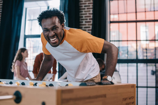 vzrušený africký americký podnikatel při pohledu na kameru při hraní stolního fotbalu v kanceláři - Fotografie, Obrázek