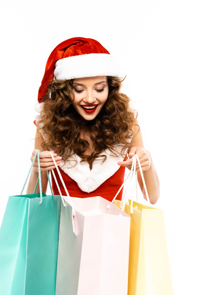 excited curly girl in santa costume looking into shopping bags, isolated on white - Fotó, kép