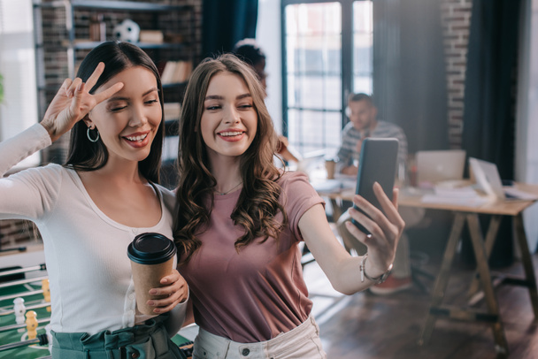 young, cheerful businesswoman taking selfie on smartphone with cheerful colleague holding coffee to go and showing victory gesture - Photo, Image