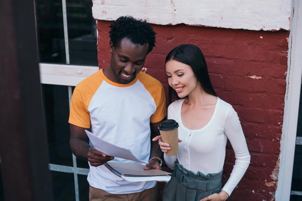 cheerful african american businessman showing documents to attractive colleague holding coffee to go  - Foto, afbeelding