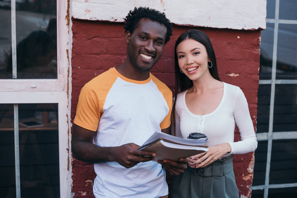 cheerful african american businessman with documents and businesswoman with coffee to go smiling at camera - Photo, image