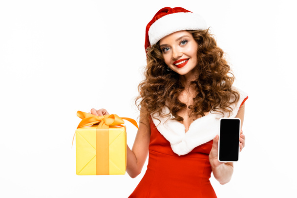 ελκυστικό χαμογελαστό κορίτσι με κοστούμι Santa κρατώντας δώρο Χριστουγέννων και smartphone με λευκή οθόνη, απομονώνονται σε λευκό - Φωτογραφία, εικόνα