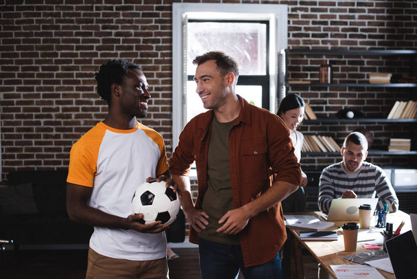 smiling african american businessman holding soccer ball near cheerful colleague standing with hand on hip - Foto, Imagem