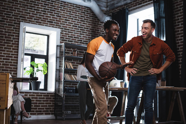 alegre afroamericano hombre de negocios celebración de baloncesto mientras habla con colega de pie con las manos en la cadera
 - Foto, Imagen