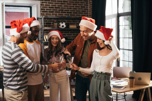 empresários multiculturais alegres em chapéus de Papai Noel clinking taças de champanhe no escritório
 - Foto, Imagem