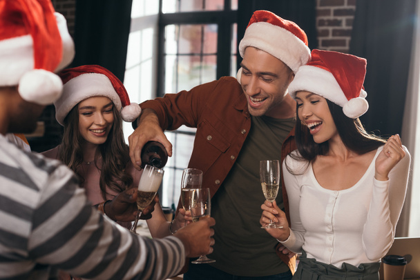 cheerful businessman pouring champagne in glasses of happy colleagues in santa hats - Фото, зображення