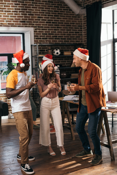 cheerful multicultural businesspeople in santa hats talking while holding champagne glasses in office - Φωτογραφία, εικόνα