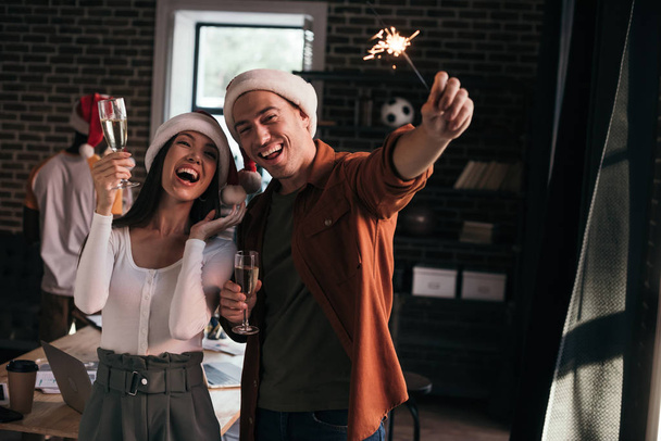Χαρούμενοι επιχειρηματίες με καπέλα santa κοιτάζοντας sparkler κρατώντας ποτήρια σαμπάνιας - Φωτογραφία, εικόνα