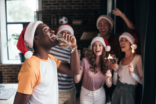 cheerful african american businessman drinking champagne while standing near multicultural colleagues in santa hats - Фото, изображение