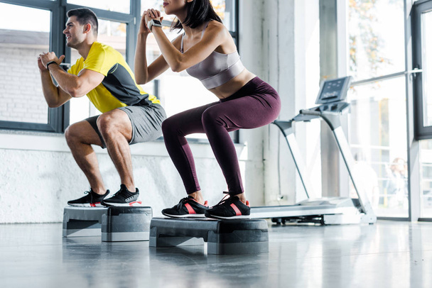 cropped view of sportsman and sportswoman doing squat on step platforms in sports center - Photo, Image