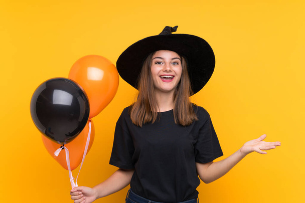 Молодая ведьма держит черные и оранжевые воздушные шары для Хэллоуинских вечеринок с шокированным выражением лица
 - Фото, изображение