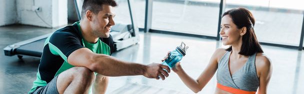 panoramic shot of smiling sportsman giving sports bottle to sportswoman in sports center - Photo, Image
