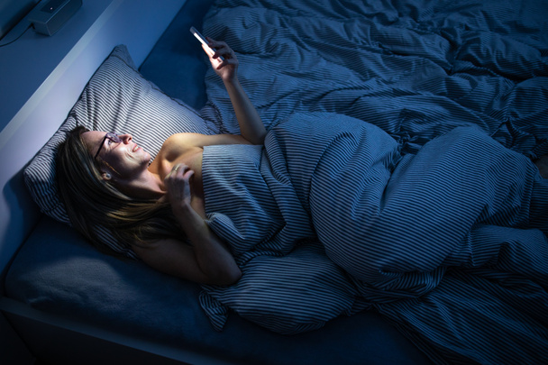 かなり中年の女性がベッドで携帯電話を使って - 写真・画像