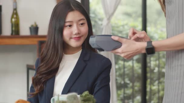 A fiatal ázsiai üzletasszonyok érintésmentesen fizetnek a kávézóban. Ázsiai boldog lány barista pincér viselni szürke kötény gazdaság hitelkártya-olvasó gép az ügyfél számára a mobiltelefon szkennelés fizetni a kávézóban. - Felvétel, videó