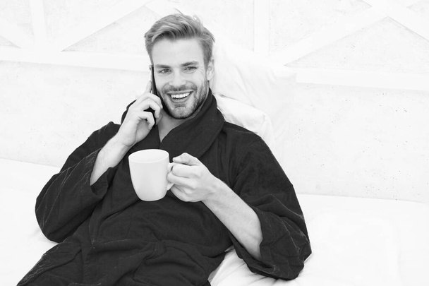 Small talk over his favorite drink. Happy man talking on phone with cup of hot drink in bed. Bearded handsome guy enjoying his drink break in bedroom. Starting his day with tasty drink - Фото, зображення