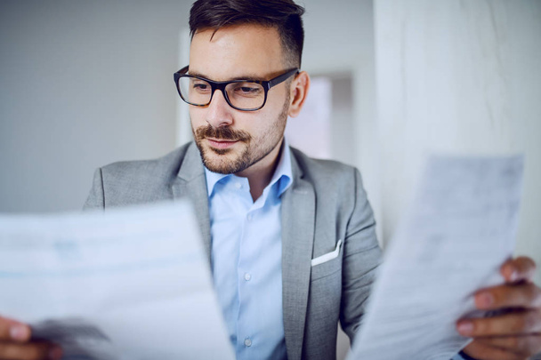 Head shot of hardworking sophisticated caucasian businessman in suit and with eyeglasses looking at documents while sitting in his office. - Foto, afbeelding