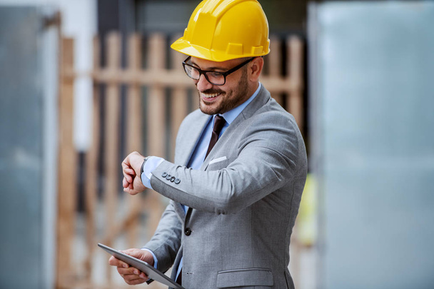 Atraente elegante arquiteto caucasiano sorridente de terno, com óculos e capacete na cabeça em pé no local de construção e segurando tablet enquanto olha para relógio de pulso
. - Foto, Imagem
