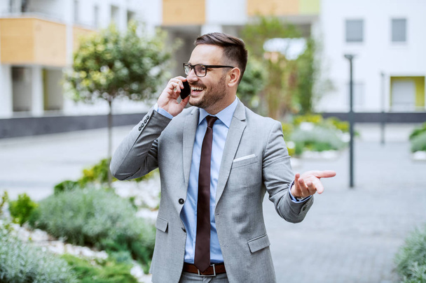 Empresário elegante caucasiano sorridente atraente em terno e com óculos falando no telefone inteligente enquanto está de pé no parque
. - Foto, Imagem