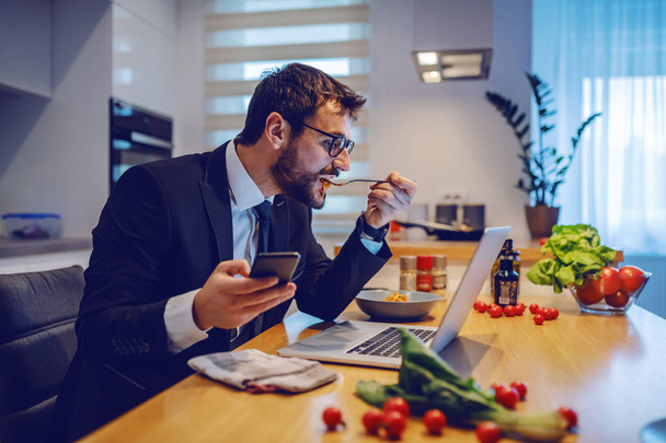 Vista lateral do atraente homem de negócios caucasiano segurando telefone inteligente, olhando para laptop e comer deliciosa refeição. Na mesa de jantar estão legumes, laptop e especiarias
. - Foto, Imagem