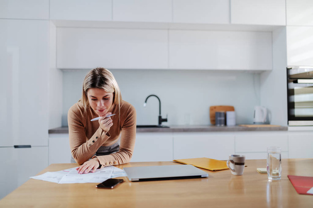 Cute caucasian blonde woman in sweater leaning on dining table and calculating bills. In hand is pen. Apartment interior. - Photo, Image