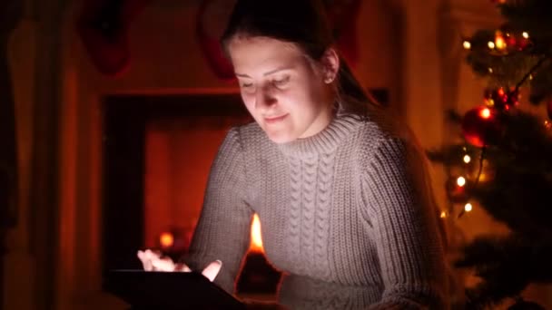 4k βίντεο της νεαρής γυναίκας με ψηφιακή ταμπλέτα κάθεται τη νύχτα δίπλα στο λαμπερό χριστουγεννιάτικο δέντρο - Πλάνα, βίντεο