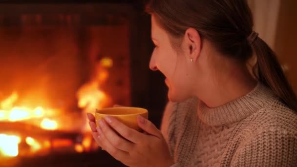 Portrait of smiling young woman looking on the fire in firepalce and drinking hot tea at night - Footage, Video