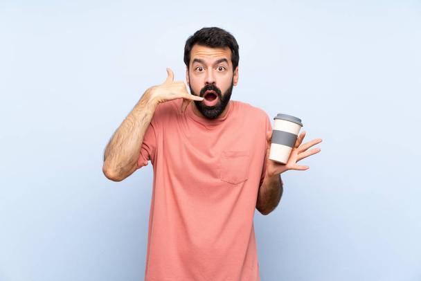 Mladý muž s plnovousem drží odnést kávu přes izolované modré pozadí dělat telefonní gesto a pochybnosti - Fotografie, Obrázek
