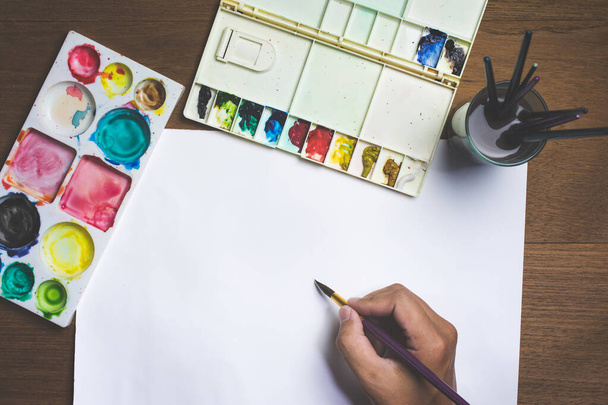 Homme main tenant pinceau de peinture sur papier blanc avec aquarelle colorée et équipement sur fond en bois
. - Photo, image
