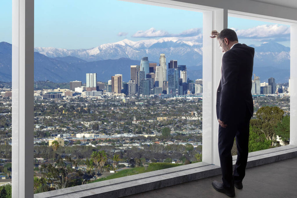 ロサンゼルスのダウンタウンの景色を眺めるオフィスのビジネスマン。男はカリフォルニアで働いている上司や地域マネージャーのように見えます.背景は冬の雪のサンガブリエル山脈を示しています.  - 写真・画像