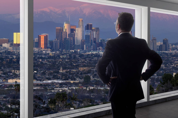 Biznesmen w garniturze, patrzący przez okno w biurze na budynki w centrum Los Angeles. Ten człowiek wygląda jak polityk jak burmistrz, architekt lub deweloper pracujący w La. - Zdjęcie, obraz