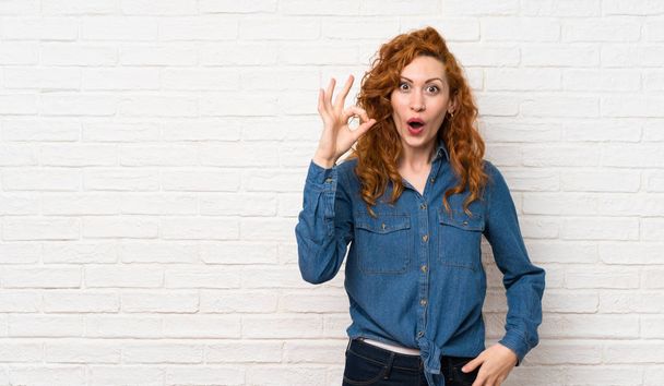 Redhead woman over white brick wall surprised and showing ok sign - Photo, Image