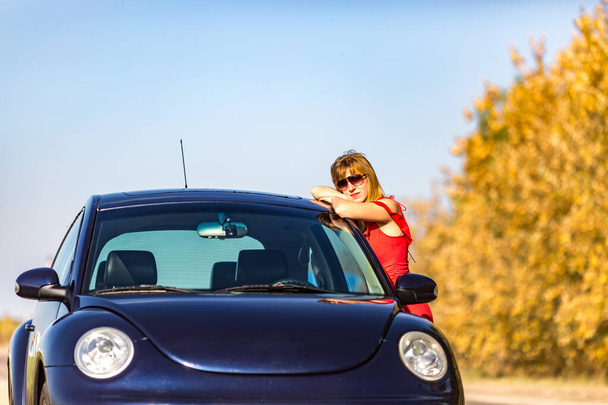Молода щаслива модель в червоному платті в сонячних окулярах біля її машини. - Фото, зображення