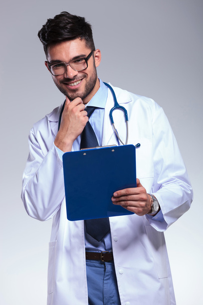 young doctor with clipboard touches his chin and smiles - Fotoğraf, Görsel