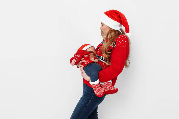 Young mother in santa hat with little baby in santa costume standing on white background - Фото, зображення