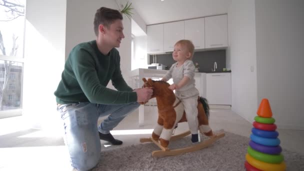 glückliche Kindheit, junger Vater spielt mit netten kleinen Jungen auf Plüschpferden und schaukelt zu Hause in der Küche - Filmmaterial, Video