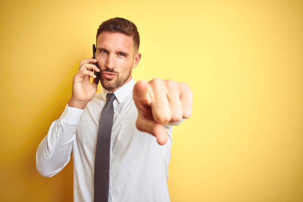 Young handsome business man talking on the phone over yellow isolated background pointing with finger to the camera and to you, hand sign, positive and confident gesture from the front - Zdjęcie, obraz