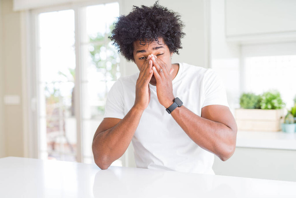 Jovem afro-americano vestindo camiseta branca casual sentado em casa esfregando os olhos para fadiga e dor de cabeça, sonolento e expressão cansada. Problema de visão
 - Foto, Imagem