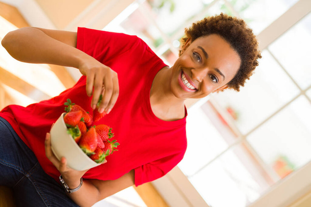 Beautiful young african woman with afro hair eating fresh strawberries sitting on the floor - Фото, зображення