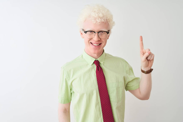 Молодой бизнесмен-альбинос в рубашке и галстуке стоит на изолированном белом фоне и показывает пальцем номер один, улыбаясь уверенно и счастливо
. - Фото, изображение
