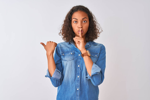 Young brazilian woman wearing denim shirt standing over isolated white background asking to be quiet with finger on lips pointing with hand to the side. Silence and secret concept. - Foto, immagini