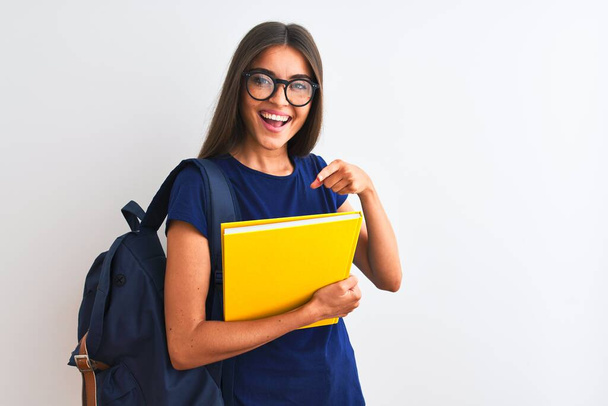 Jovem estudante mulher usando óculos mochila segurando livro sobre fundo branco isolado muito feliz apontando com a mão e o dedo
 - Foto, Imagem