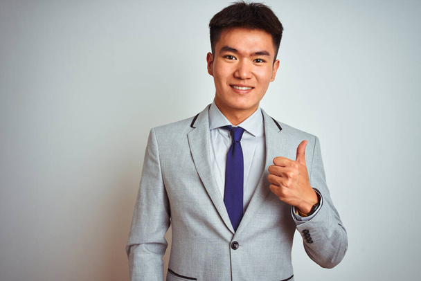 Homme d'affaires chinois asiatique vêtu d'une veste grise et cravate debout sur un fond blanc isolé faisant des pouces heureux geste avec la main. Approuver l'expression en regardant la caméra montrant le succès
. - Photo, image