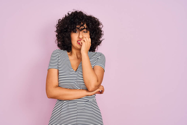 Young arab woman with curly hair wearing striped dress over isolated pink background looking stressed and nervous with hands on mouth biting nails. Anxiety problem. - 写真・画像